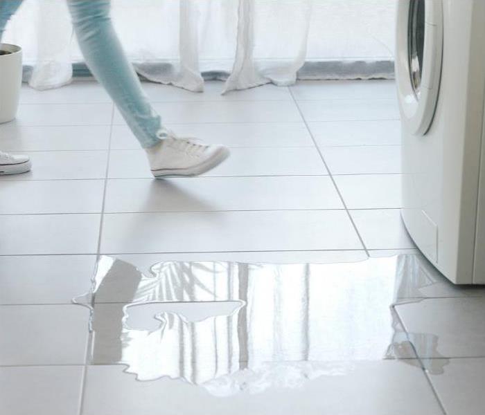 A person wearing white shoes walks on a tile floor next to a washing machine. There is a puddle of water on the floor.