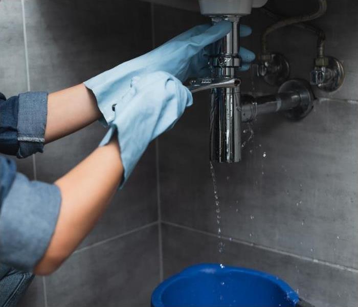 Hands wearing gloves holding a wrench, reaching under a sink where the sink is dripping