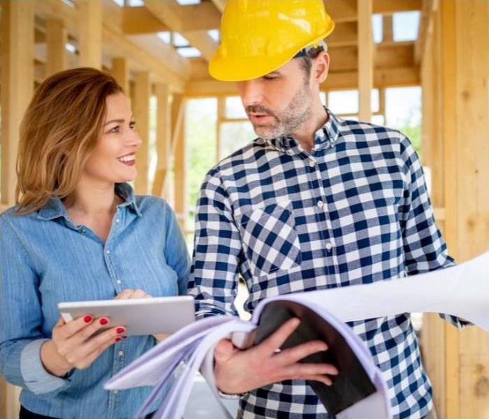 A woman and man, wearing a hard hat, look at blueprints at a construction site.